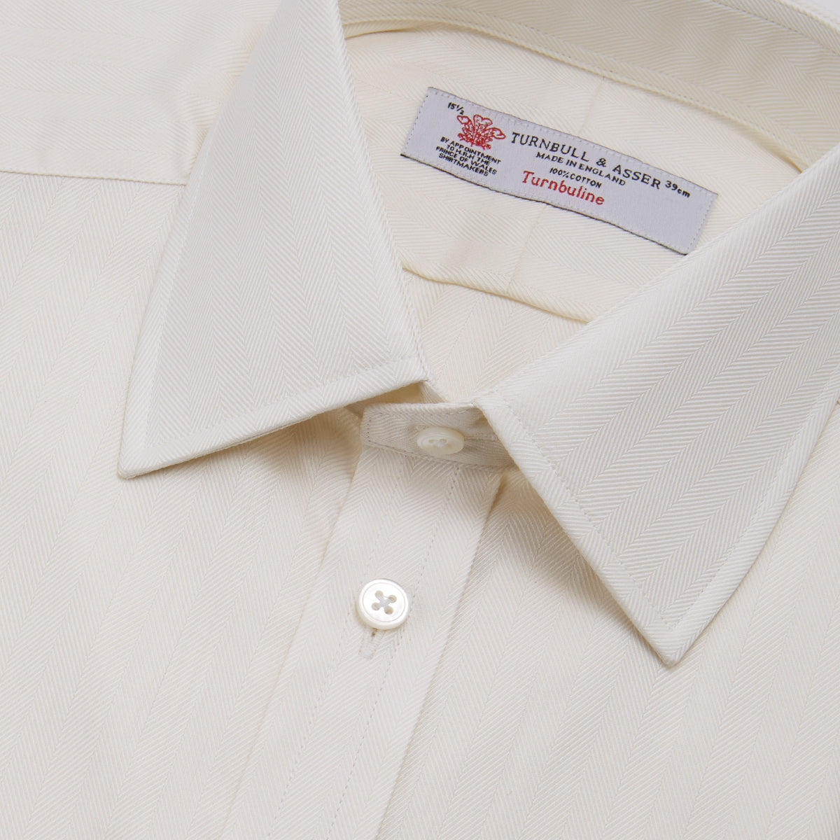 View 5 of product The Turnbull & Asser Cream Herringbone Superfine Cotton Shirt features a T&A collar, double cuffs, and a subtle pattern. It is neatly folded with white buttons and displayed on a plain white background.