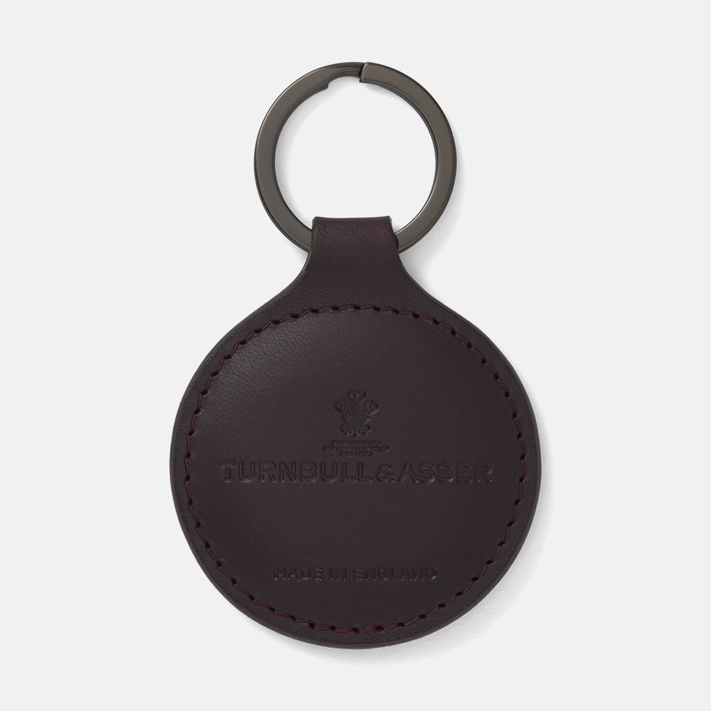 The Violla Leather Key Fob by Turnbull & Asser is a round, dark brown leather accessory with embossed branding, visible edge stitching, and a metal loop at the top. Made in England, it combines elegance and function for your keys.