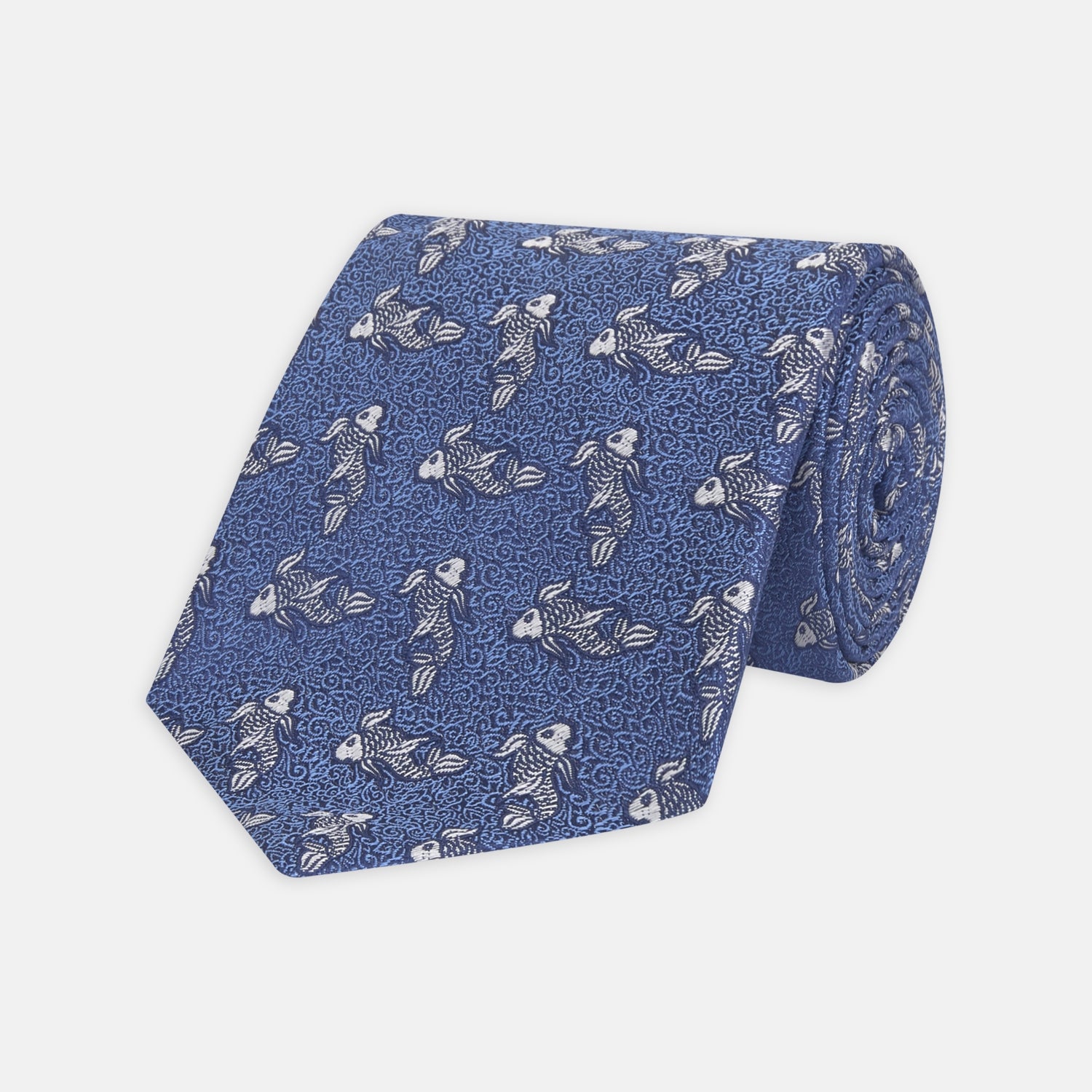 View 1 of product The Blue Carp Silk Tie by Turnbull & Asser features a blue silk base with white and dark blue koi fish among light blue swirls. The tie is displayed rolled up, pointed end forward.