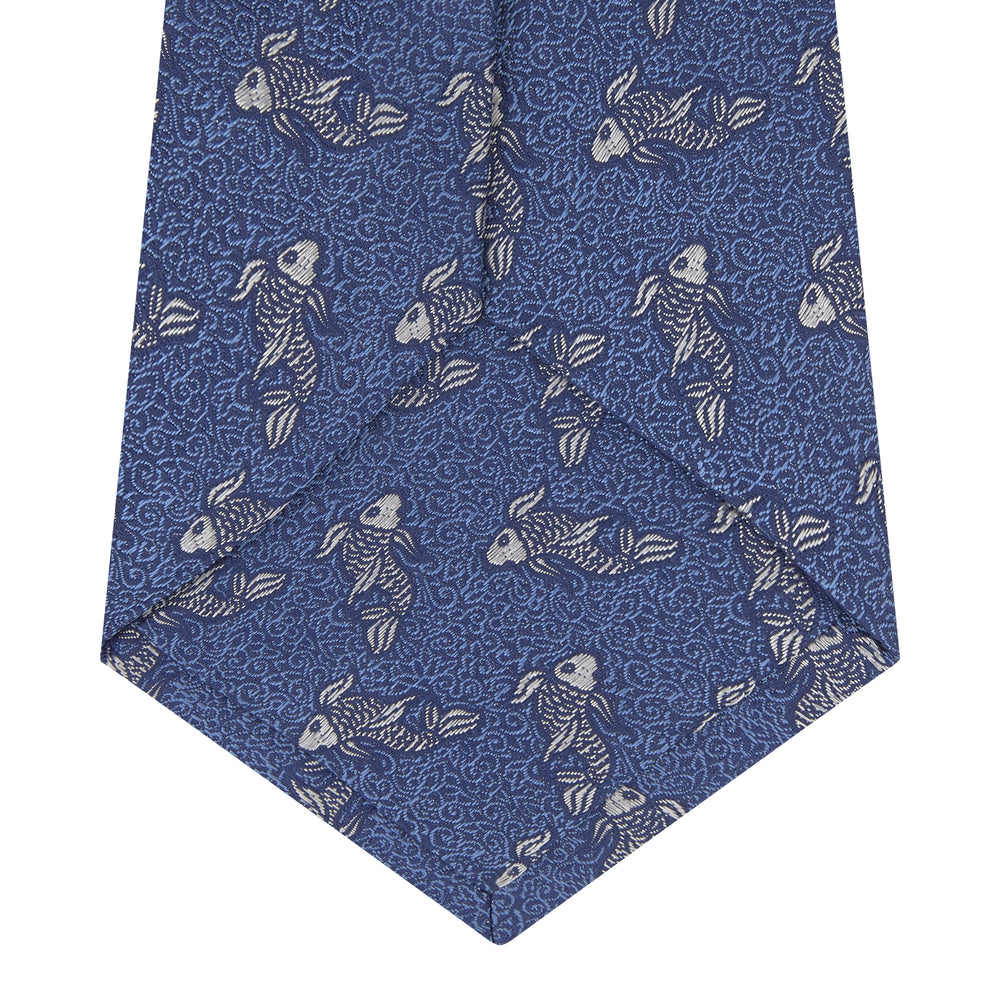 The Blue Carp Silk Tie by Turnbull & Asser features a blue silk base with white and dark blue koi fish among light blue swirls. The tie is displayed rolled up, pointed end forward.