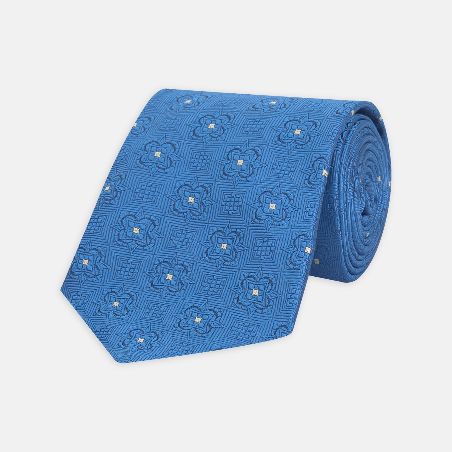 View 1 of product The Turnbull & Asser Blue Flower Mosaic Silk Tie features a blue silk fabric with a detailed geometric and floral design, accented in lighter blue and white, displayed on a white background.